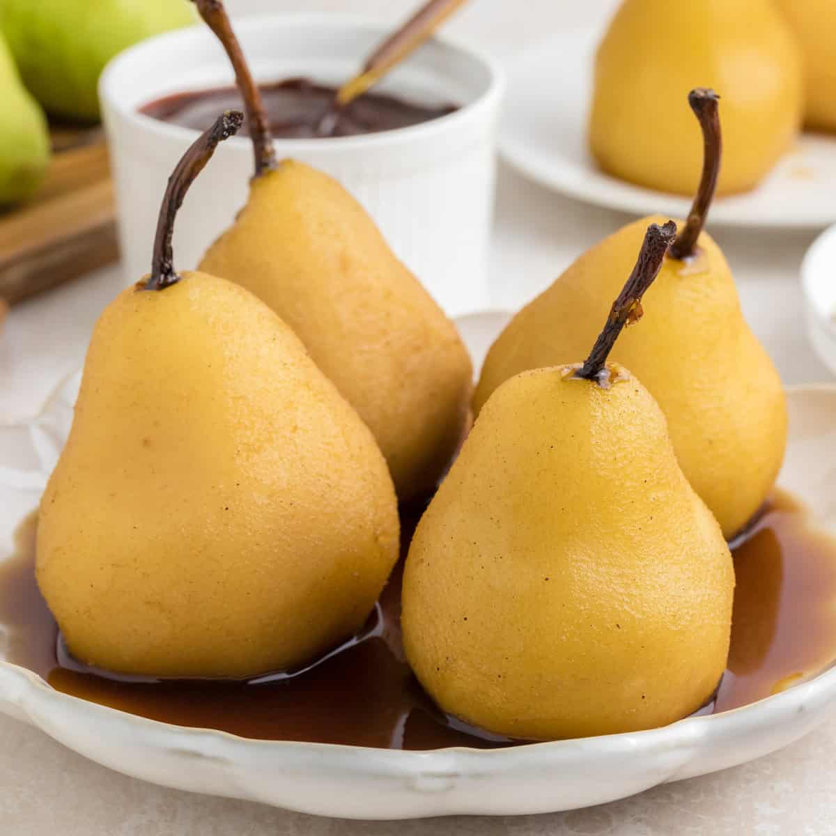Four poached pears sit on a white plate in a pool of brown sauce, with a small bowl of sauce and additional pears visible in the background.