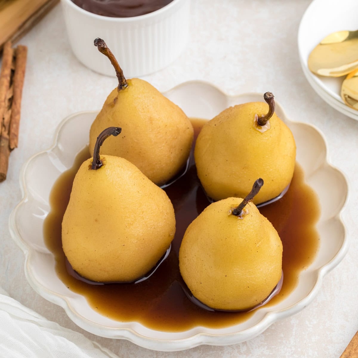 Four poached pears are served on a white plate with a brown sauce, with cinnamon sticks and a small bowl of sauce in the background.
