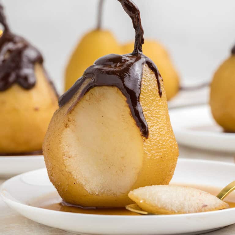 A poached pear with chocolate sauce drizzled on top sits on a plate, with a spoonful of the pear placed in front.