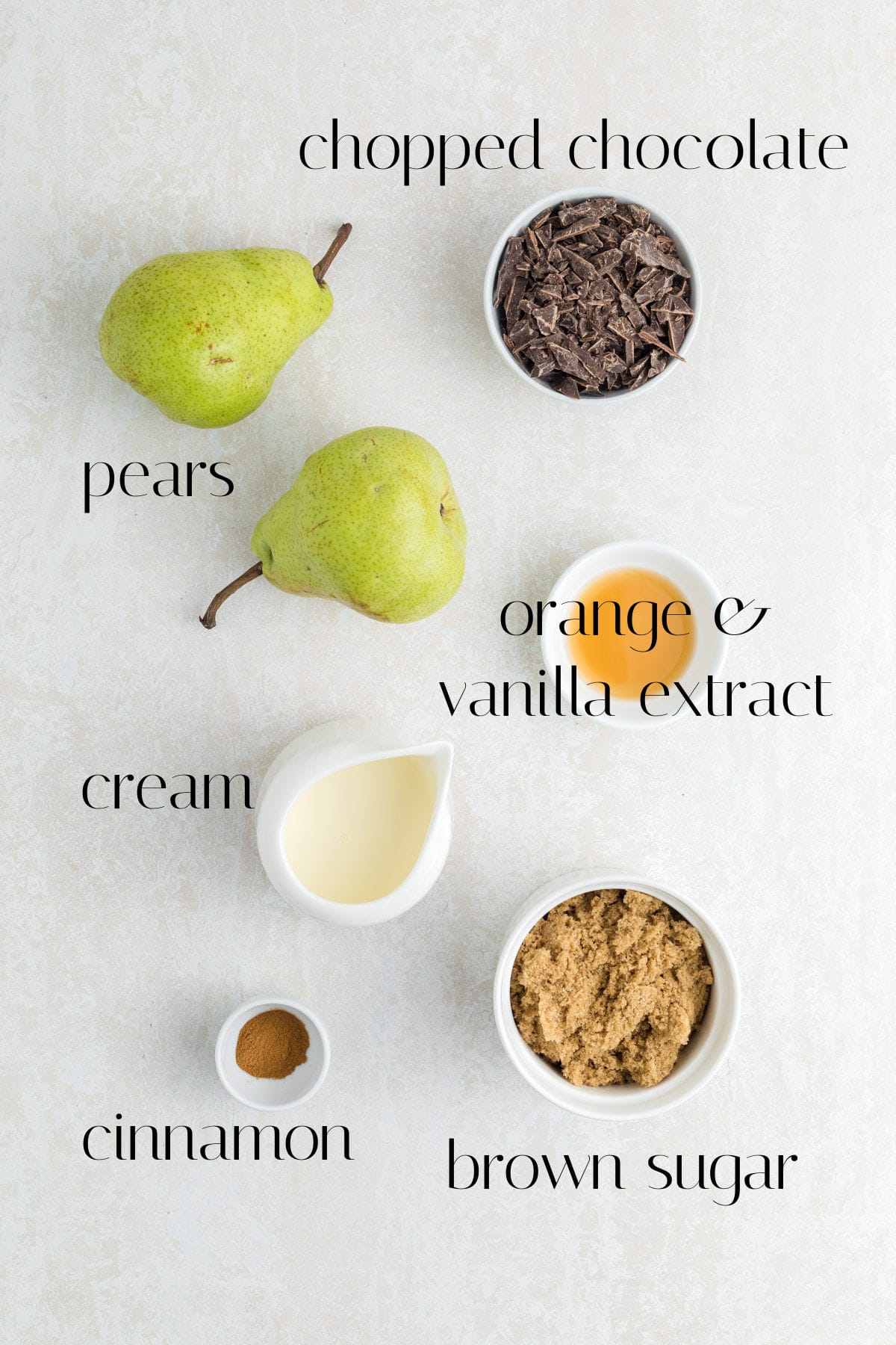 Two green pears, a bowl of chopped chocolate, a small bowl of brown sugar, a small jug of cream, a bowl of orange and vanilla extract, and a pinch of cinnamon, all labeled on a light surface.