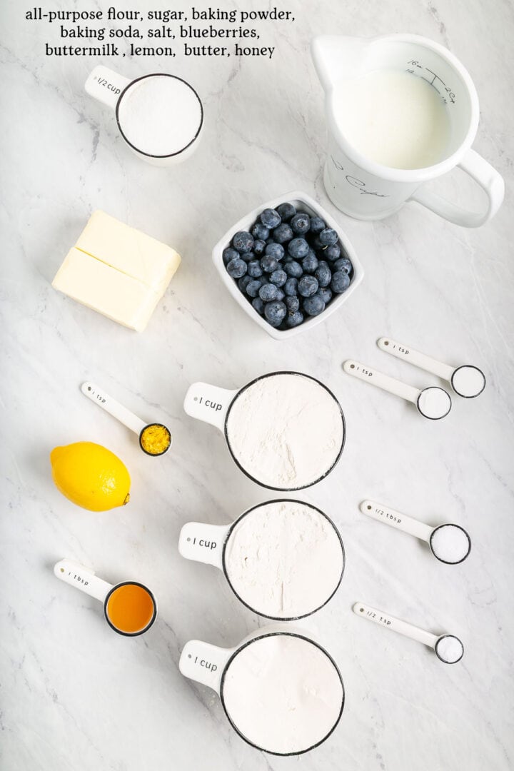 Overhead view of baking ingredients on a white surface: flour, sugar, baking powder, baking soda, salt, buttermilk, blueberries, butter, honey, a lemon, and measuring spoons and cups filled with ingredients.