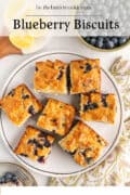 A plate of square blueberry biscuits is arranged on a table with a small bowl of fresh blueberries, lemon slices on a cutting board, and a fork on a small plate nearby. The text Blueberry Biscuits is at the top.