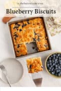 Overhead view of golden blueberry biscuits cut into squares in a baking pan, one piece lifted on a spatula. Next to the pan are a bowl of fresh blueberries, a plate with a fork, and a floral napkin.