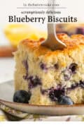A close-up of a fluffy blueberry biscuit on a plate, with fresh blueberries and a fork cutting into it. The text above reads: Scrumptiously delicious Blueberry Biscuits by thebuttercookie.com.