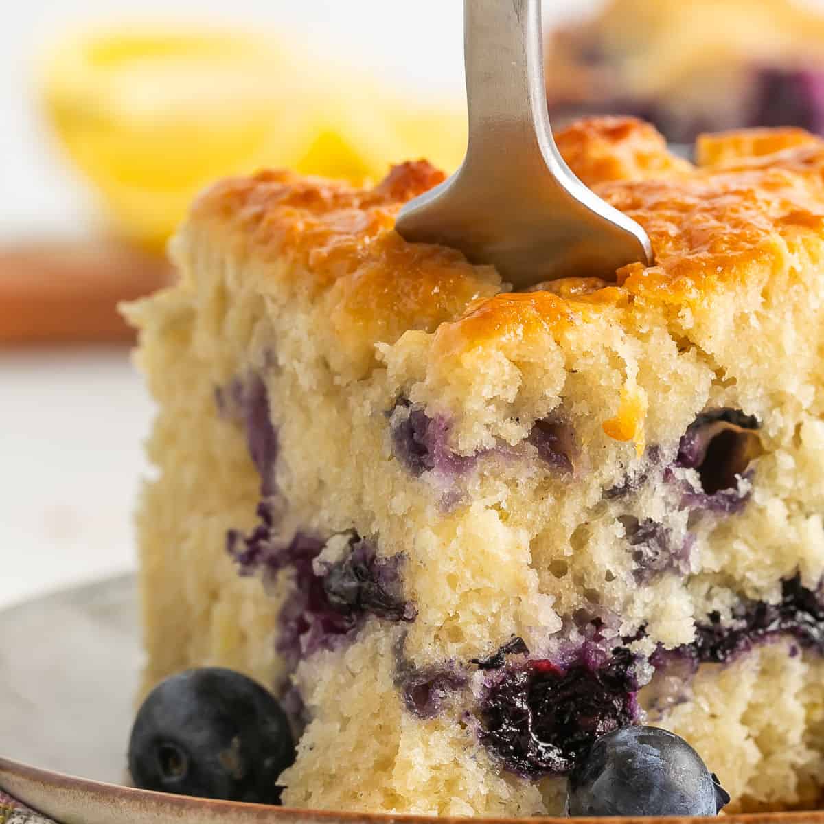 A close-up of a moist slice of blueberry lemon cake on a plate, with a fork inserted into the fluffy top and fresh blueberries scattered around. A blurred lemon is visible in the background.
