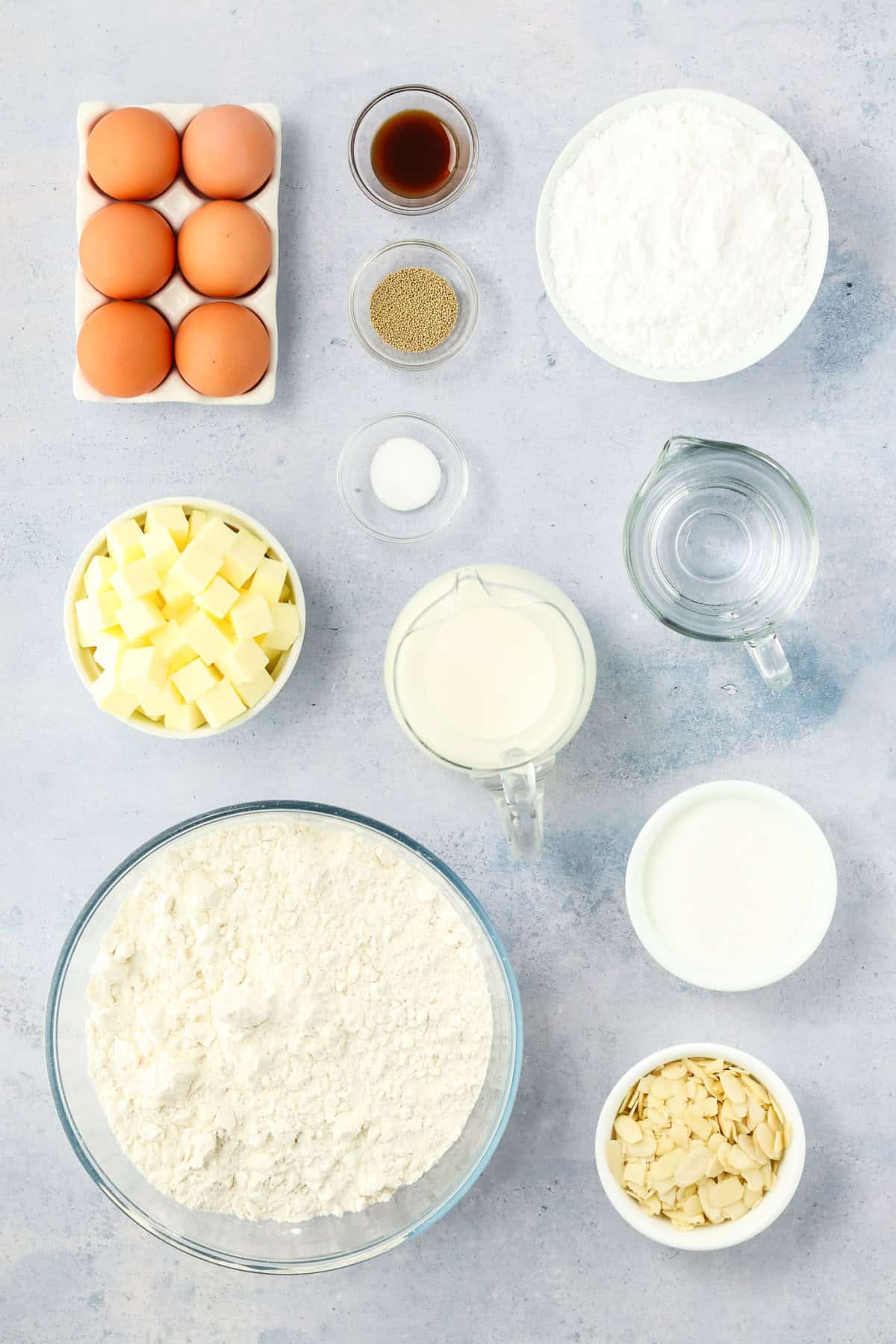 Danish Butterhorn baking ingredients arranged on a light surface, including eggs, cubes of butter, flour, sugar, milk, water, sliced almonds, vanilla, salt, and yeast in bowls and containers.