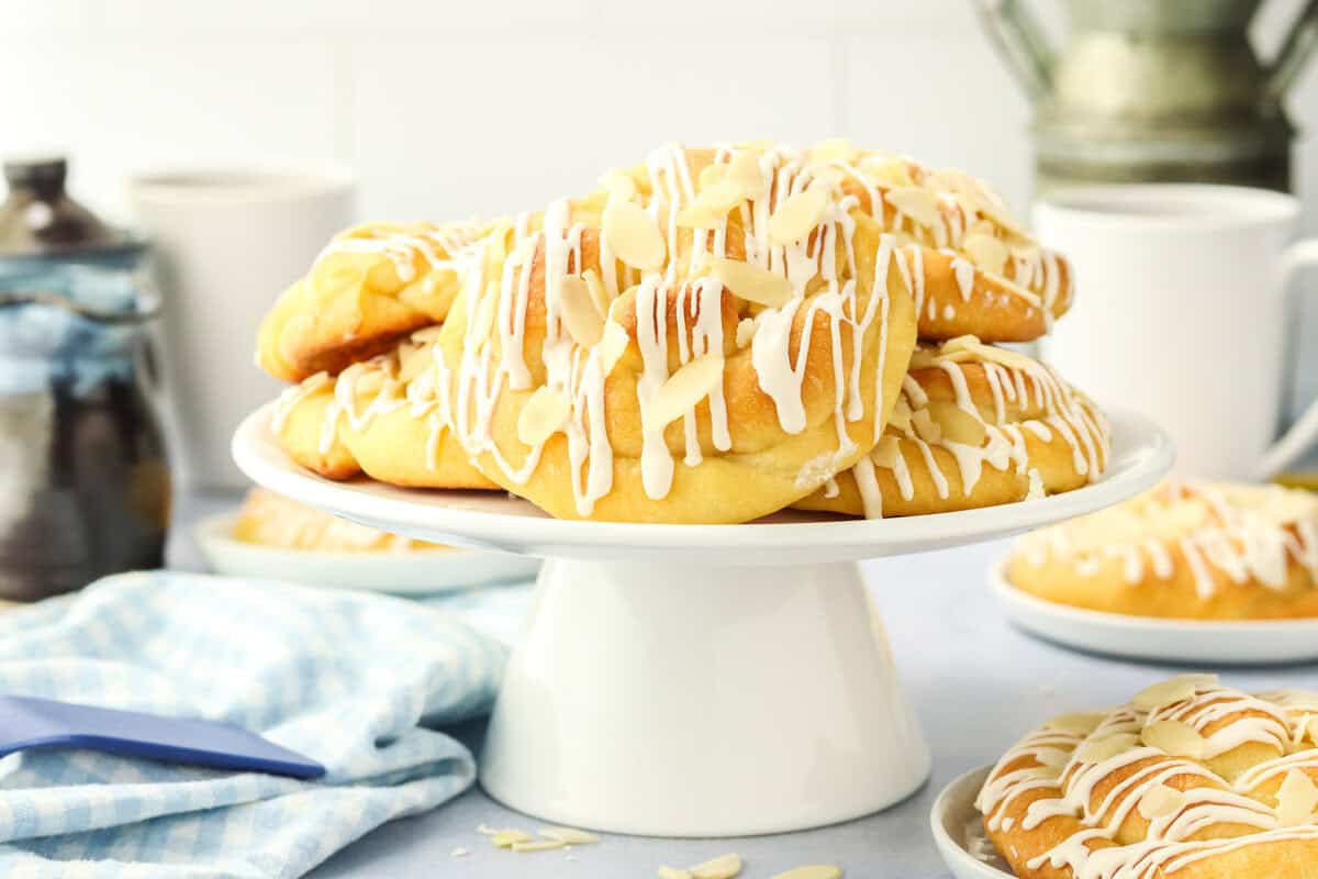 A stack of Danish Butterhorns topped with sliced almonds and white icing sits on a white cake stand. More pastries, a blue cloth, and kitchenware are visible in the background.
