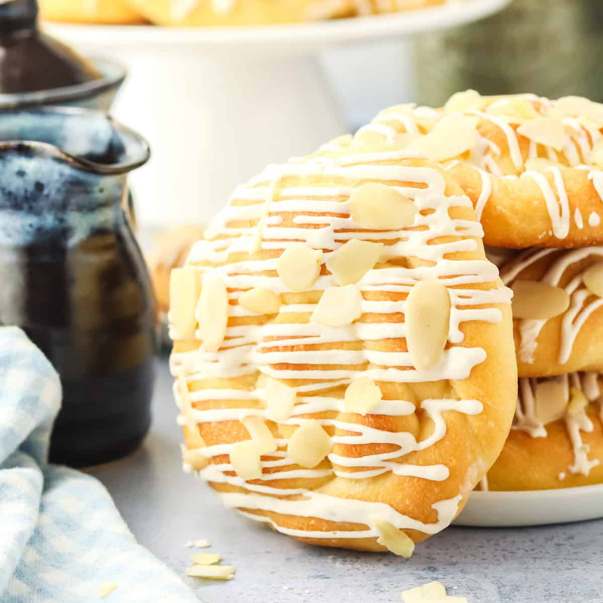 A close-up of danish butterhorns pastry drizzled with white icing and topped with sliced almonds, stacked on a white plate with a blue checkered cloth and a ceramic pitcher nearby.