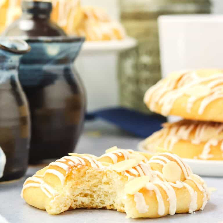 A close-up of a partially eaten pastry drizzled with white icing and topped with almond slices, with more pastries and ceramic dishes blurred in the background.
