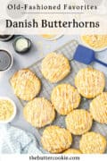 A cooling rack holds Danish Butterhorn pastries drizzled with icing. Nearby are a blue spatula, coffee cup, milk, sliced almonds, and a blue checkered cloth. The text reads: “Danish Butterhorns” and “thebuttercookie.com.”.