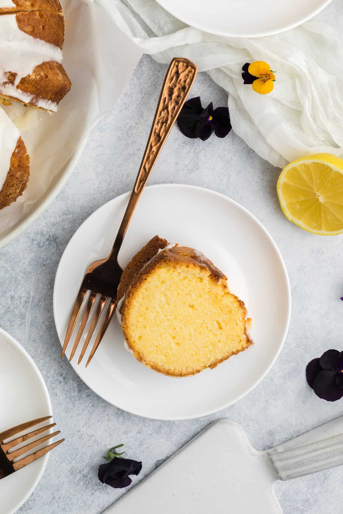 A slice of bundt cake on a white plate with a gold fork, surrounded by lemon, dark purple flowers, and a white cloth on a light surface.
