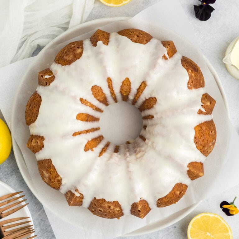 A round lemon bundt cake covered with a smooth white glaze sits on parchment paper, surrounded by lemon halves, a fork, a flower, and a white cloth, creating a bright and inviting presentation.