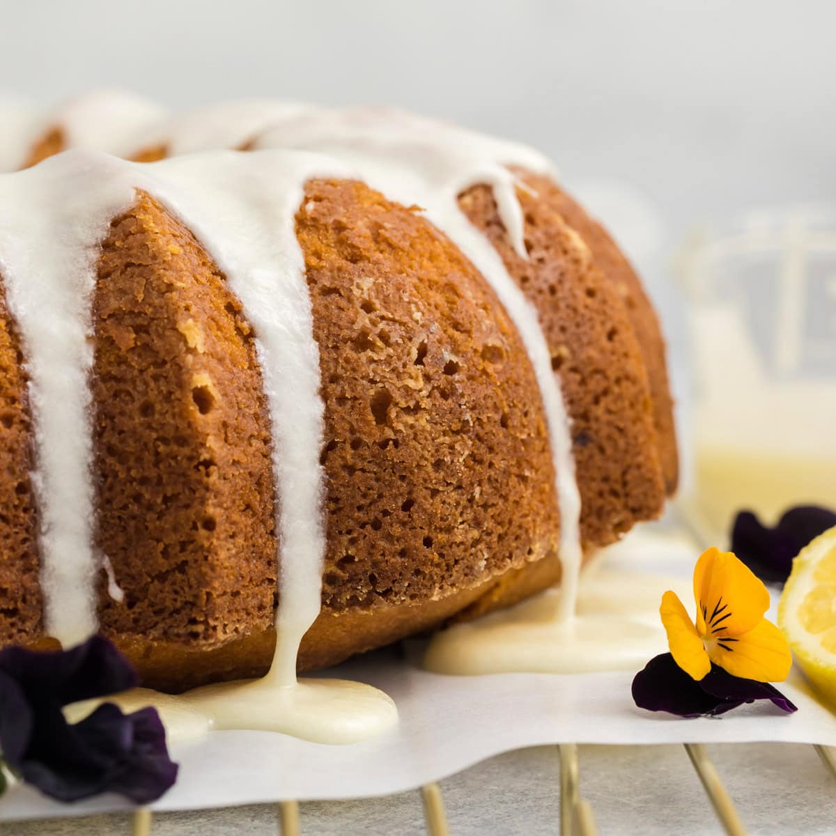 A close-up of a glazed lemon bundt cake with white icing drizzling down the sides, garnished with edible yellow and purple flowers and a lemon slice on a cooling rack.