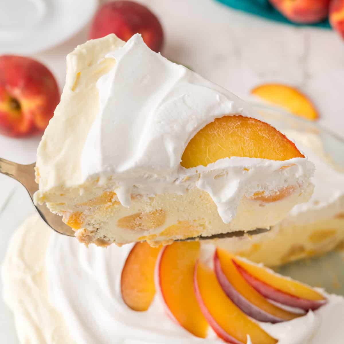 A close-up of a slice of creamy peach dessert pie with whipped topping and fresh peach slices, being lifted from the whole pie, which is garnished with more peach slices. Whole peaches are visible in the background.
