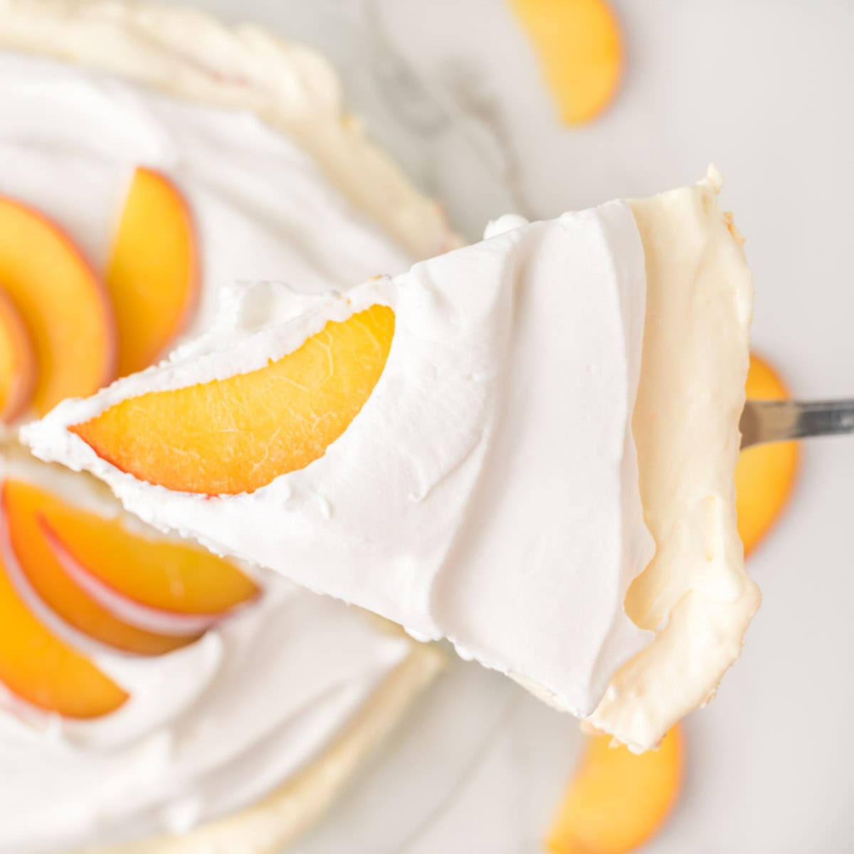 A close-up of a slice of creamy pie topped with a peach slice, being lifted from the whole pie, which is decorated with more peach slices and swirls of whipped topping.