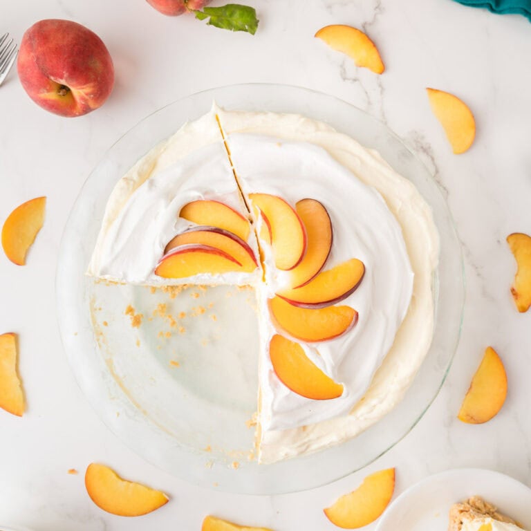 A peaches and cream jello pie topped with peach slices sits on a white surface. One slice is missing, and peach slices are scattered around the pie. A whole peach and a fork are visible nearby.