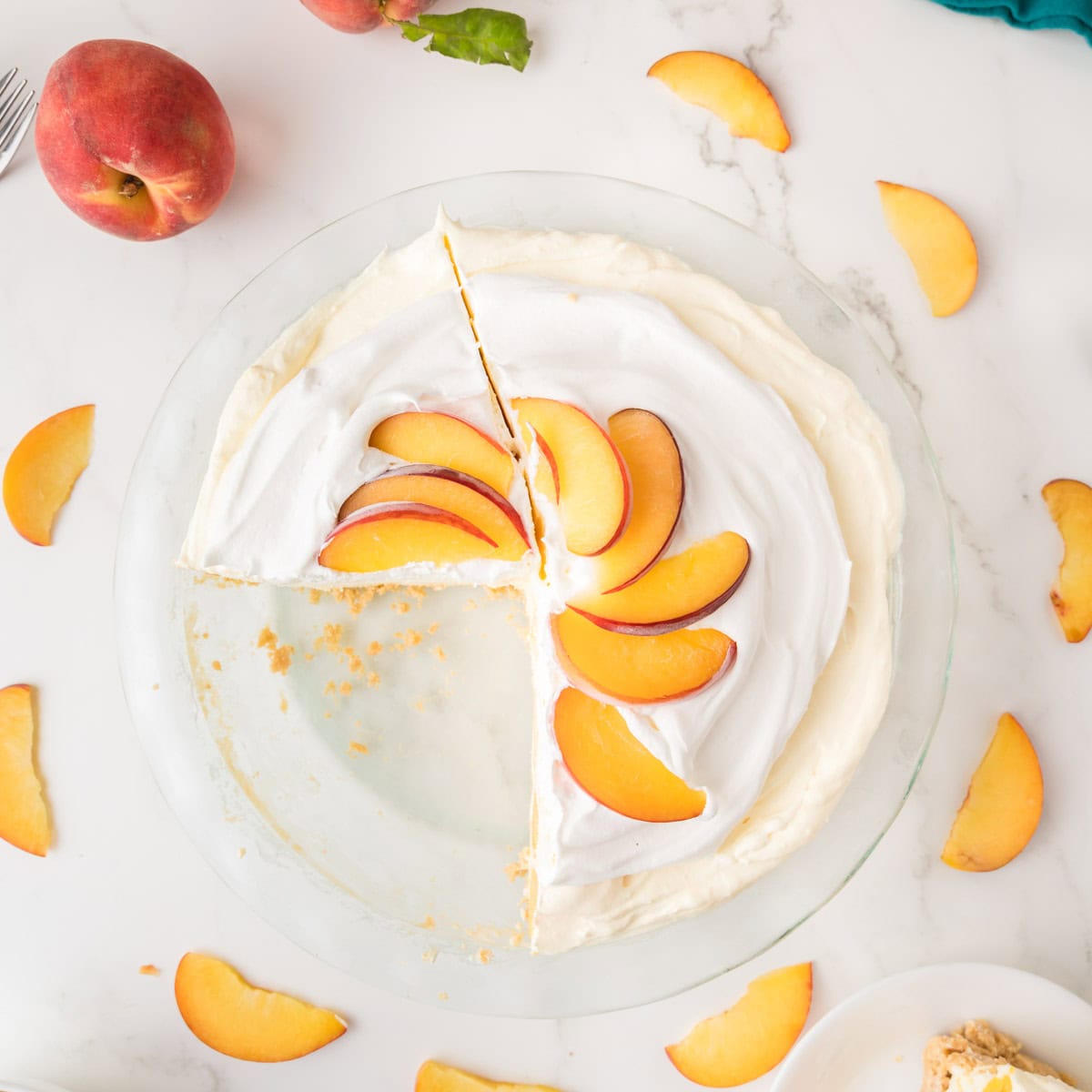 A peaches and cream jello pie topped with peach slices sits on a white surface. One slice is missing, and peach slices are scattered around the pie. A whole peach and a fork are visible nearby.