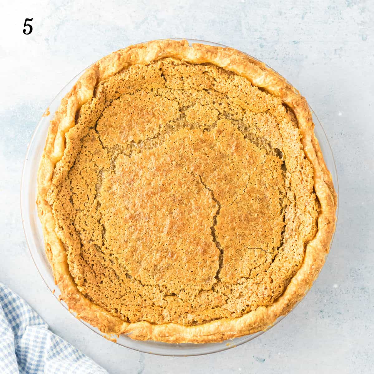 A backwoods maple pie with a cracked top sits in a glass pie dish on a light surface, with a blue and white checkered cloth at the bottom left. The number 5 is in the upper left corner.