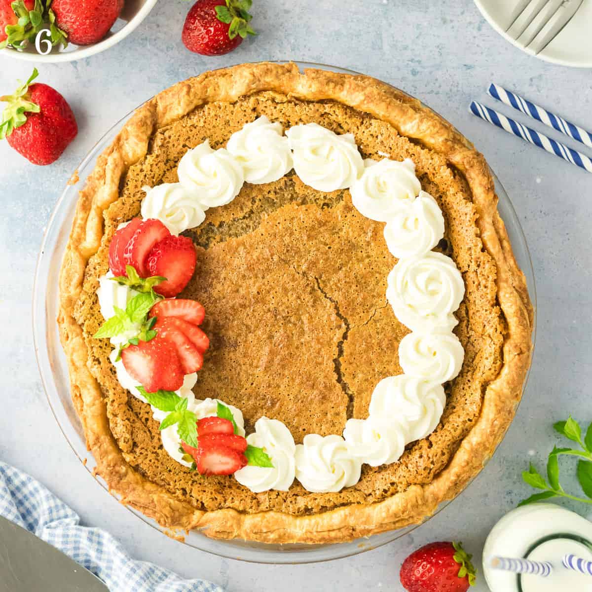 A golden-brown maple pie topped with a ring of white whipped cream, garnished with sliced strawberries and mint leaves, sits on a light blue surface surrounded by fresh strawberries and plates.
