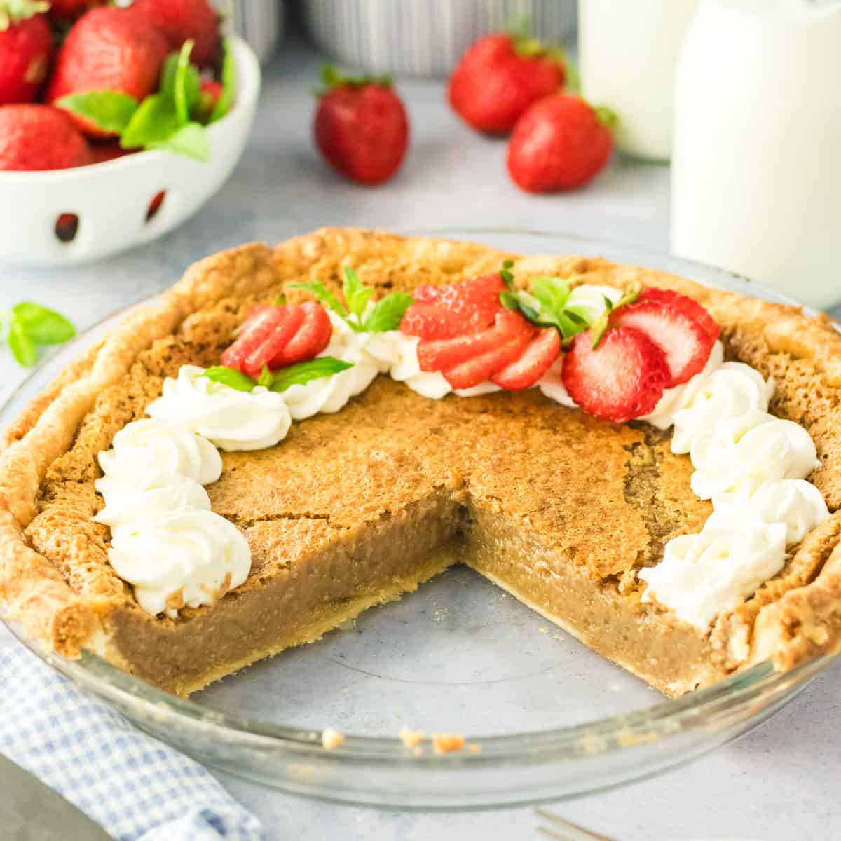 A backwoods maple pie with a golden crust and creamy filling sits on a counter, missing one slice. The pie is topped with piped whipped cream and fresh sliced strawberries, with more strawberries and bottles of milk in the background.