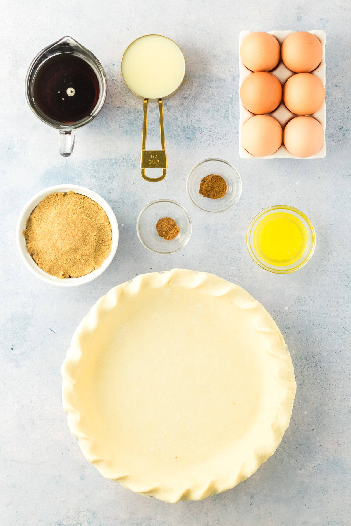 Overhead view of ingredients for a pie: a raw pie crust, six eggs, melted butter, brown sugar, ground spices, sweetened condensed milk, and dark syrup, all arranged on a light surface.