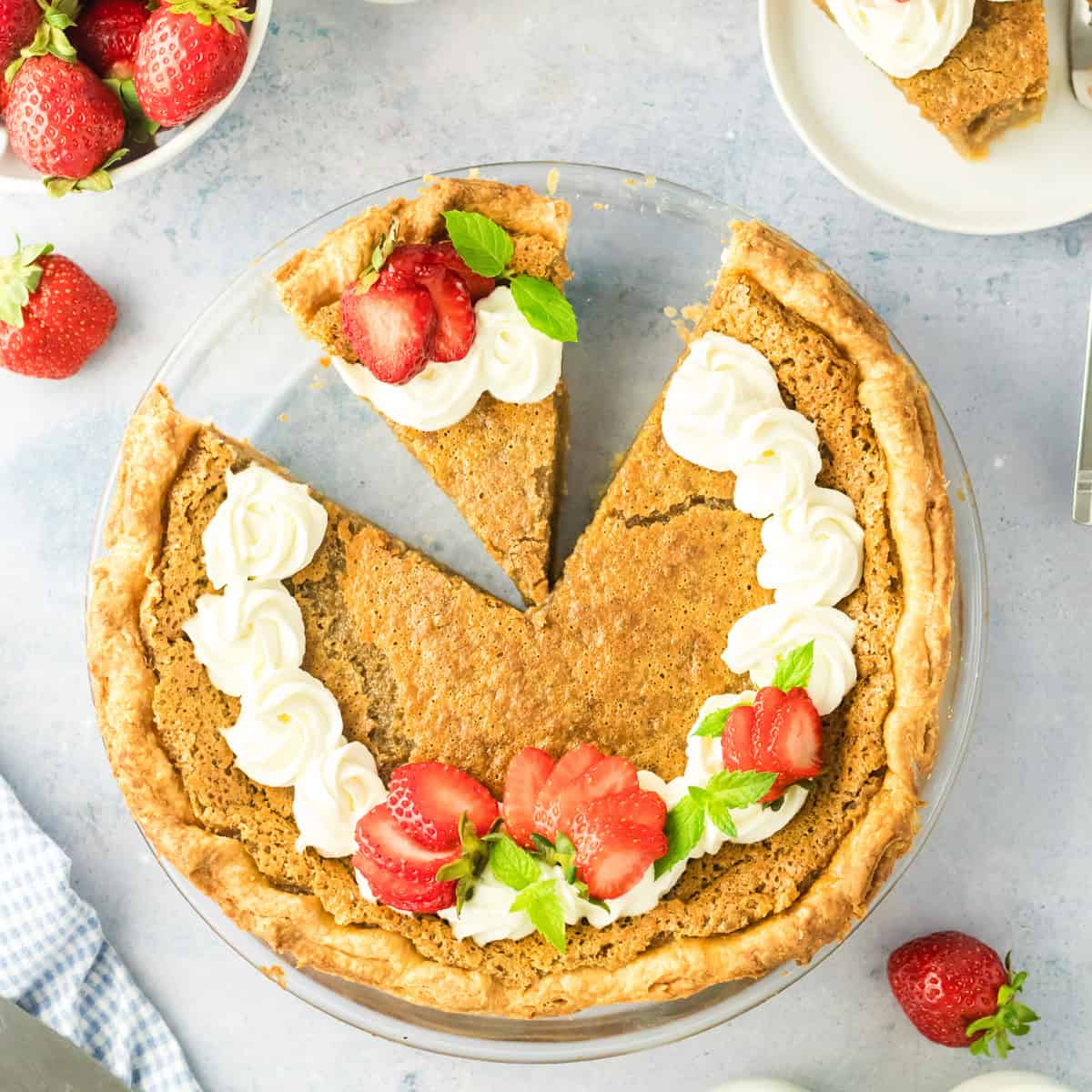 A golden-brown maple syrup pie with a slice removed, topped with swirls of whipped cream, sliced strawberries, and mint leaves. Fresh strawberries are scattered nearby on a light blue surface.