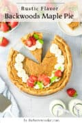 A backwoods maple pie in a glass dish, topped with whipped cream, strawberries, and mint leaves. A slice is served on a plate nearby. Fresh strawberries, milk bottles, and a pie server are also visible.