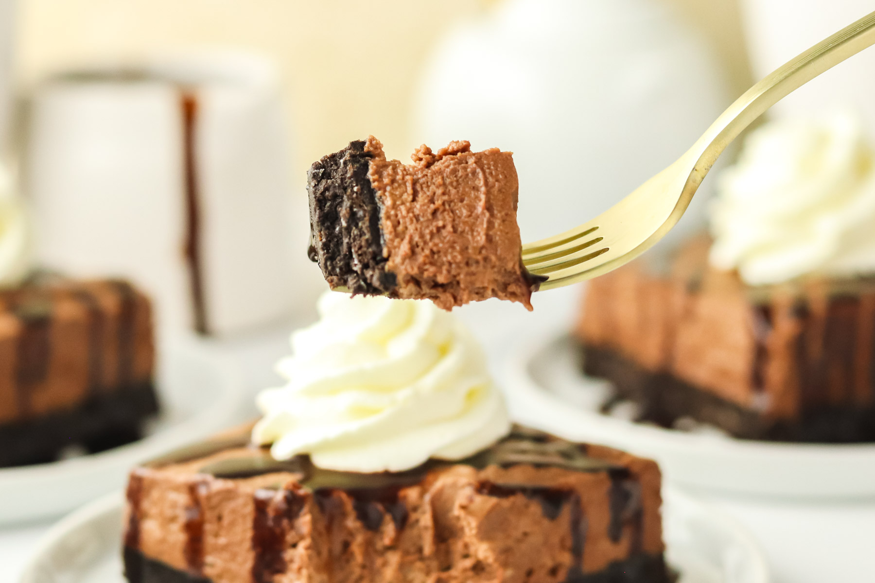 A fork holds a bite of chocolate mousse cheesecake with a chocolate crust, in front of a slice topped with a swirl of whipped cream. More cheesecake slices are blurred in the background.