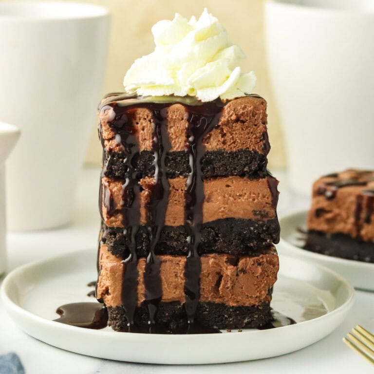 A tall slice of layered chocolate cheesecake with an Oreo crust, topped with whipped cream and drizzled with chocolate sauce, sits on a white plate. A fork rests nearby and a blurred dessert is in the background.