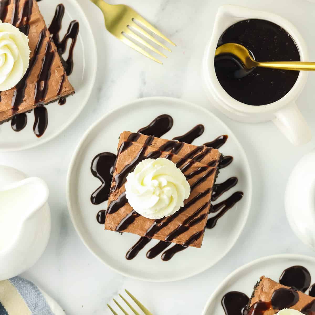 A slice of chocolate dessert topped with whipped cream and chocolate drizzle on a white plate, surrounded by gold forks, chocolate sauce in a small pitcher, and additional dessert plates.