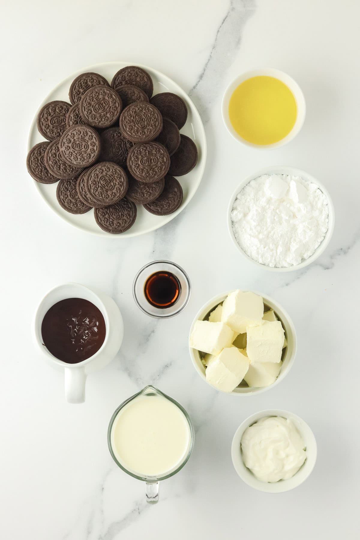A top view of ingredients on a white surface, including a plate of chocolate oreo cookies, melted butter, powdered sugar, chocolate sauce, vanilla extract, cubes of cream cheese, heavy cream, and sour cream in bowls and cups.