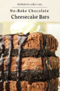 A close-up of stacked no-bake chocolate cheesecake bars with a chocolate cookie crust, topped with whipped cream and drizzled with chocolate sauce.