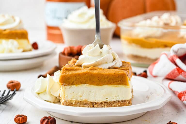 A slice of pumpkin cheesecake bars with a graham cracker crust, topped with whipped cream, sits on a white plate. A fork is inserted into the cheesecake. Pumpkin decor and more cheesecake are in the background.