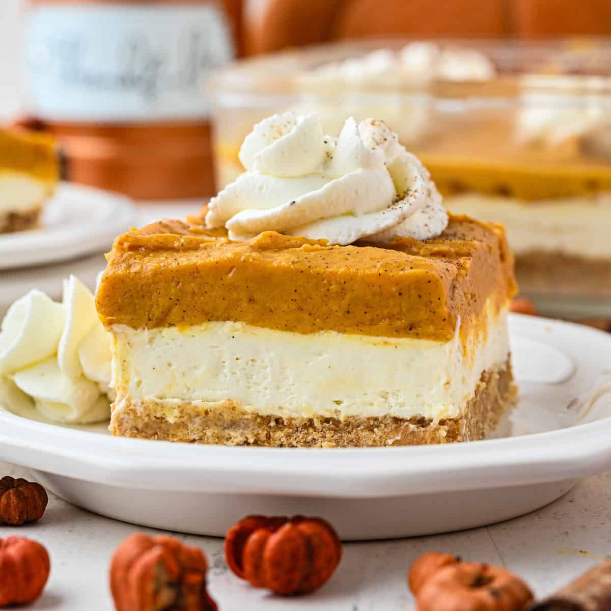 A slice of pumpkin cheesecake with a graham cracker crust, creamy middle layer, and pumpkin topping, garnished with swirls of whipped cream on a white plate, with more cheesecake and autumn decor in the background.