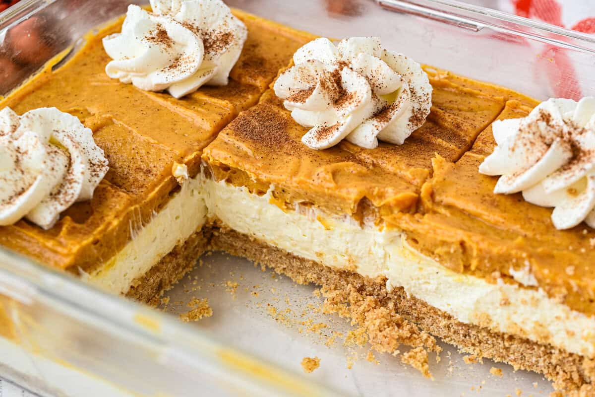 A glass baking dish with pumpkin cheesecake bars featuring a graham cracker crust, creamy cheesecake layer, pumpkin topping, whipped cream swirls, and a dusting of cinnamon. One section of the dessert has been cut out.