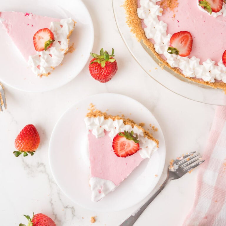 A pink strawberry cream pie topped with whipped cream and fresh strawberries is sliced and served on white plates. Whole strawberries and a fork are on a marble surface nearby.