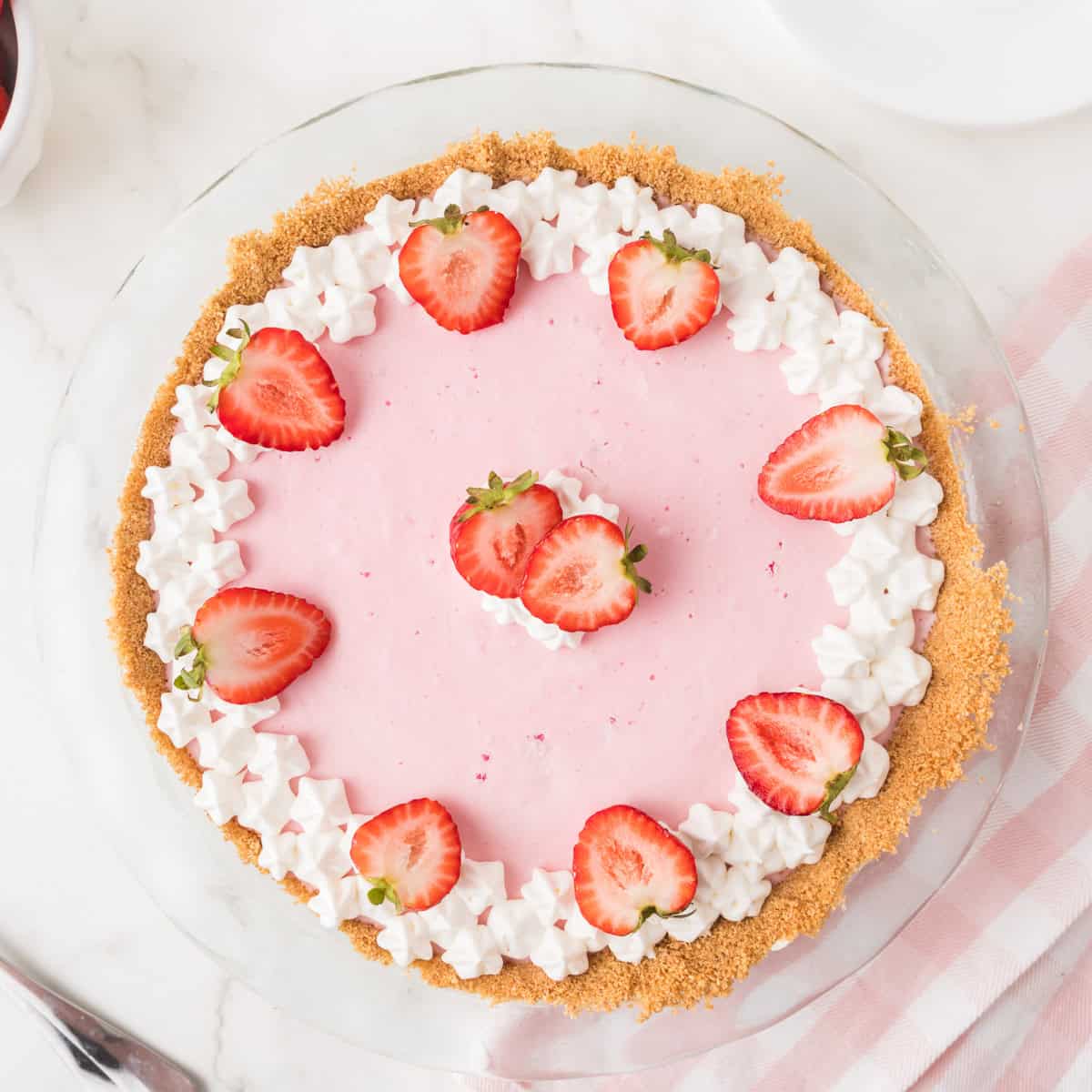 A pink cream pie in a graham cracker crust, topped with dollops of whipped cream and halved fresh strawberries, arranged in a circle on top. The pie is on a white surface with a pink striped napkin nearby.