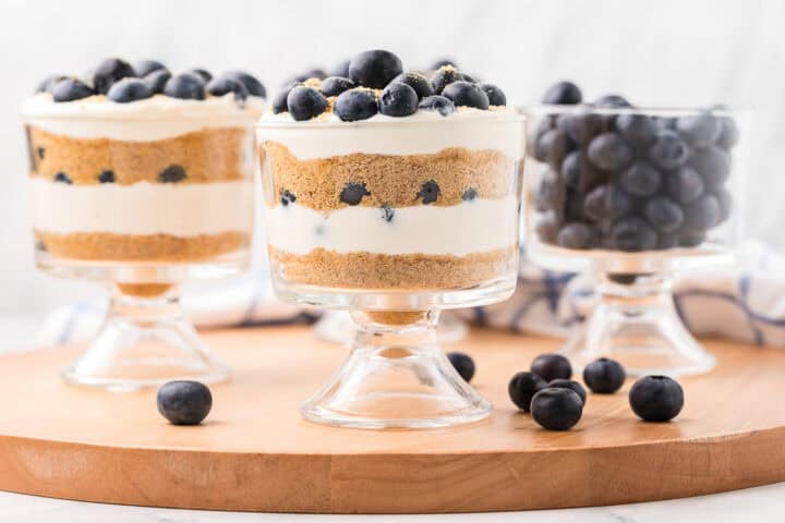 Three glass dessert cups on a wooden board—two filled with layered blueberry parfait cheesecake, graham cracker crumbs, cream, and blueberries, and one filled with fresh blueberries. Loose blueberries are scattered around.