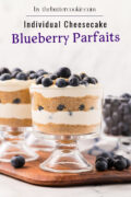 Two glass cups filled with layers of graham cracker crumbs, creamy cheesecake filling, and blueberries, topped with fresh blueberries, sit on a wooden board. A bowl of blueberries and scattered berries are in the background.