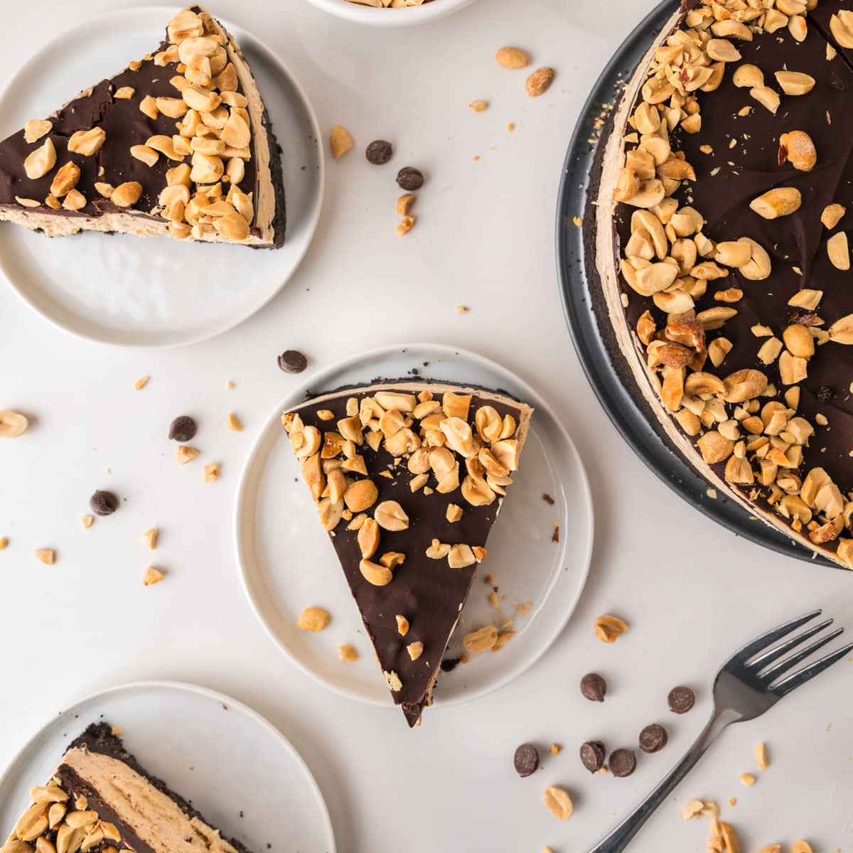 A sliced chocolate peanut butter pie topped with chopped peanuts is served on white plates. Some slices are arranged around the whole pie on a white surface, with scattered peanuts and chocolate chips. A fork is placed nearby.