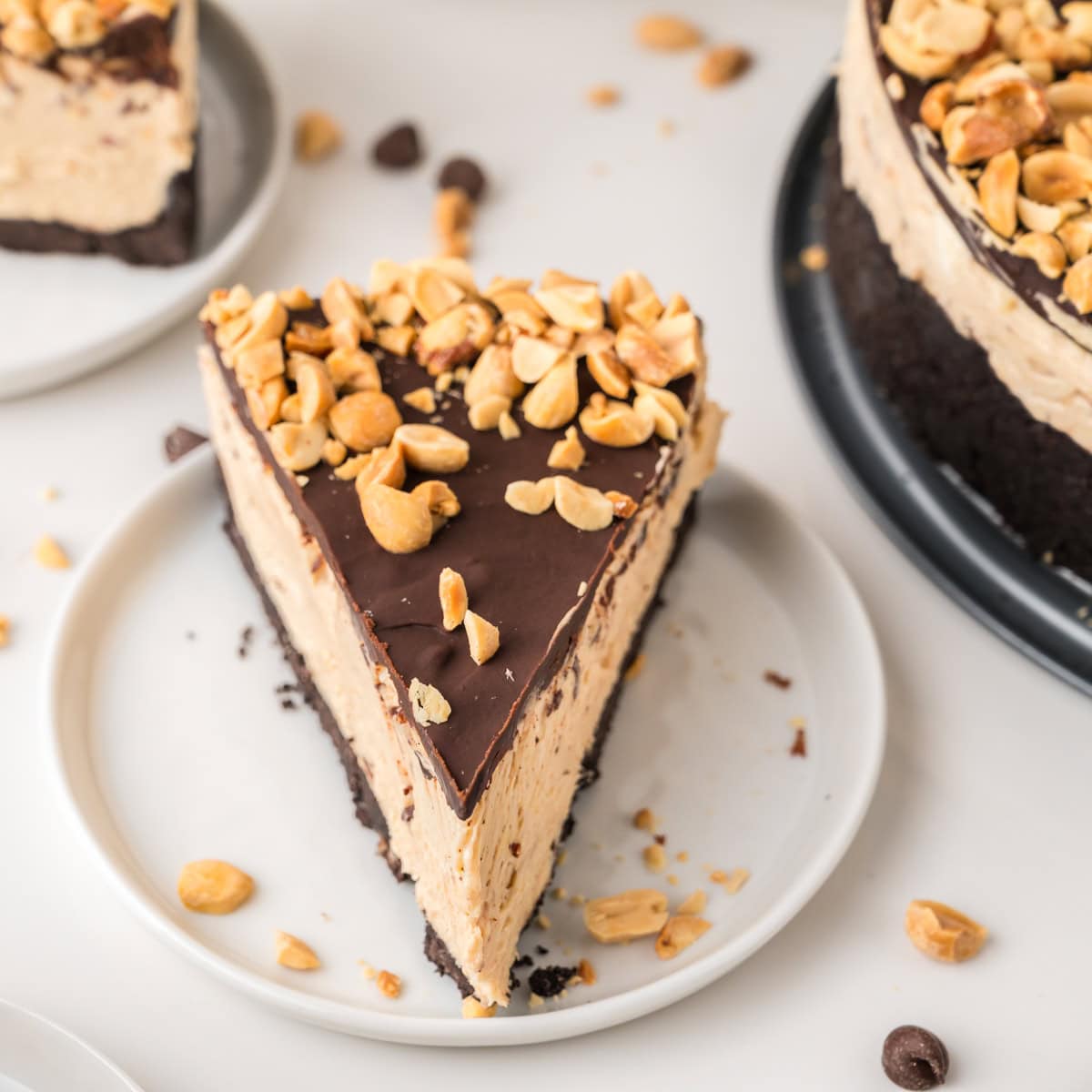 A slice of peanut butter cheesecake with a chocolate topping and chopped peanuts on a white plate, with crumbs and peanuts scattered around.