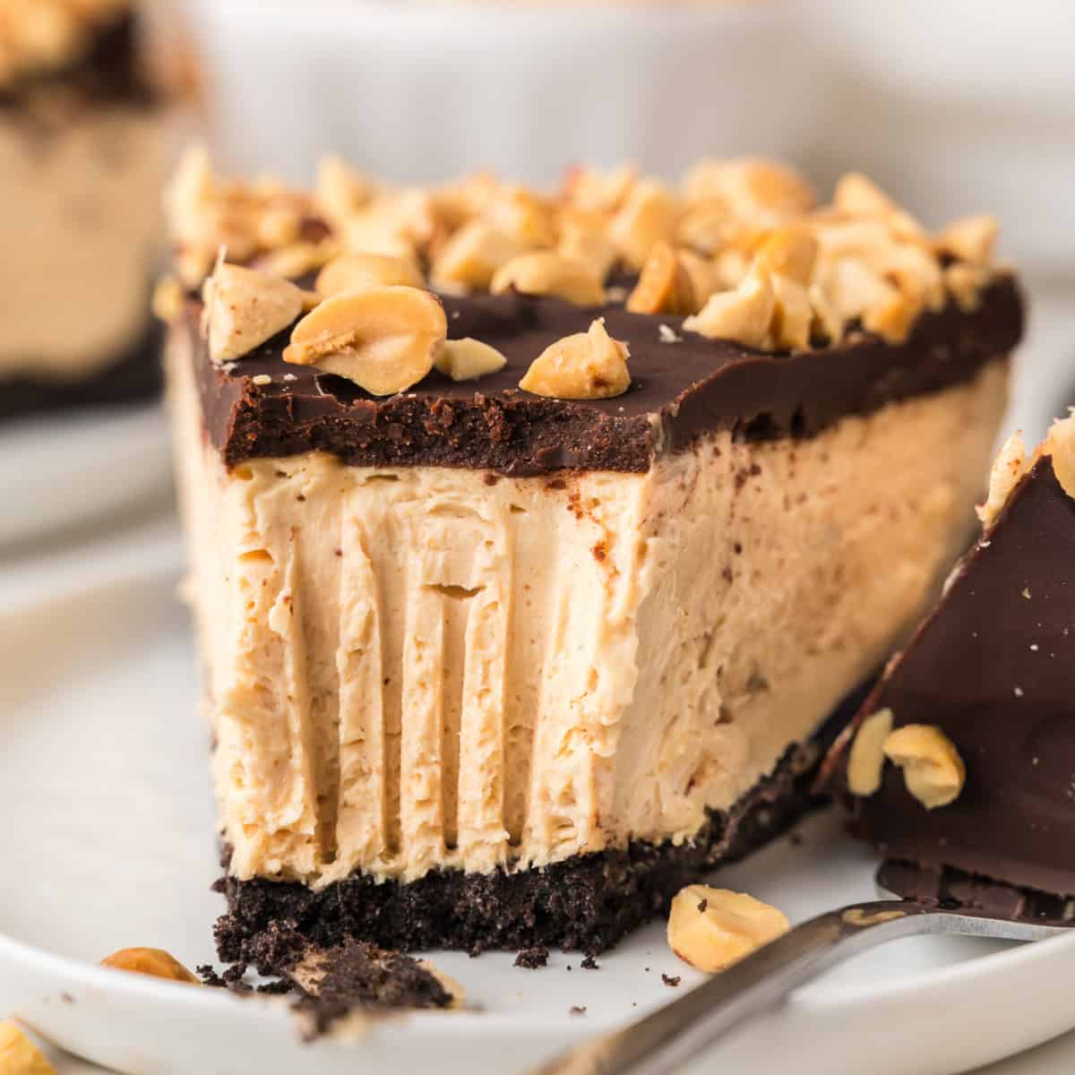 A slice of peanut butter cheesecake topped with chopped peanuts and a layer of chocolate sits on a white plate. A fork marks a bite taken from the creamy cheesecake, revealing its rich texture.