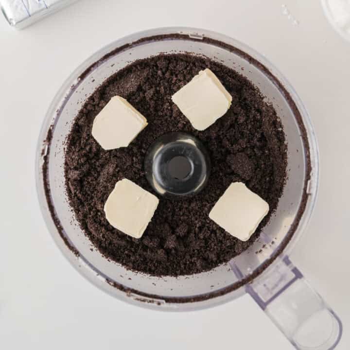 A food processor containing crushed chocolate cookies and five cubes of butter, ready to be mixed together.