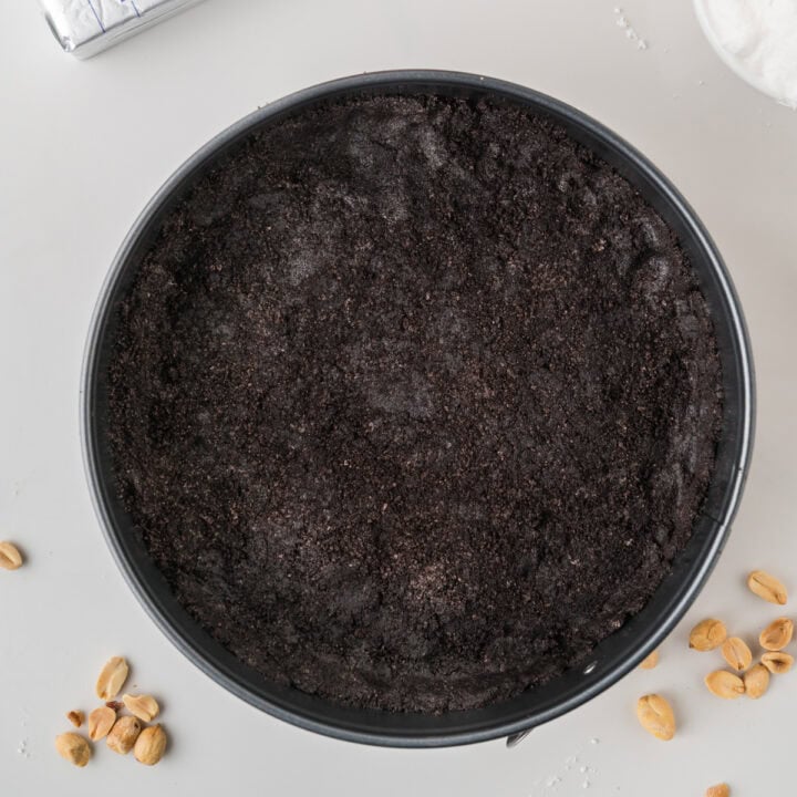 A round baking pan filled with a dark, crumbly cookie crust sits on a light surface. Scattered peanuts are around the pan, and a partially visible bowl of whipped topping is in the corner.