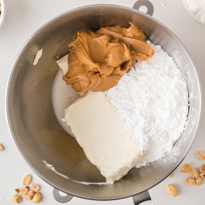 A mixing bowl containing peanut butter, powdered sugar, and a block of cream cheese, with scattered peanuts on the white surface nearby.