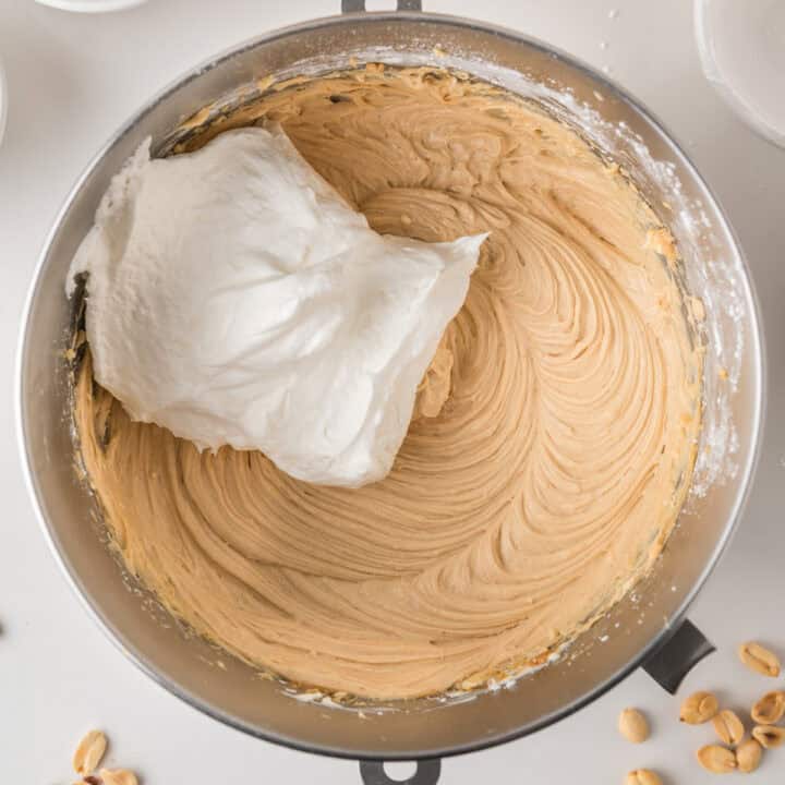 A metal mixing bowl contains creamy peanut butter batter with a large dollop of fluffy whipped topping on one side, ready to be folded in. Scattered peanuts are visible on the white counter nearby.