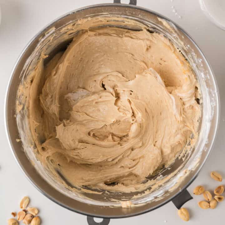 A mixing bowl filled with creamy, light brown peanut butter frosting, with a few peanuts scattered on the white surface nearby.