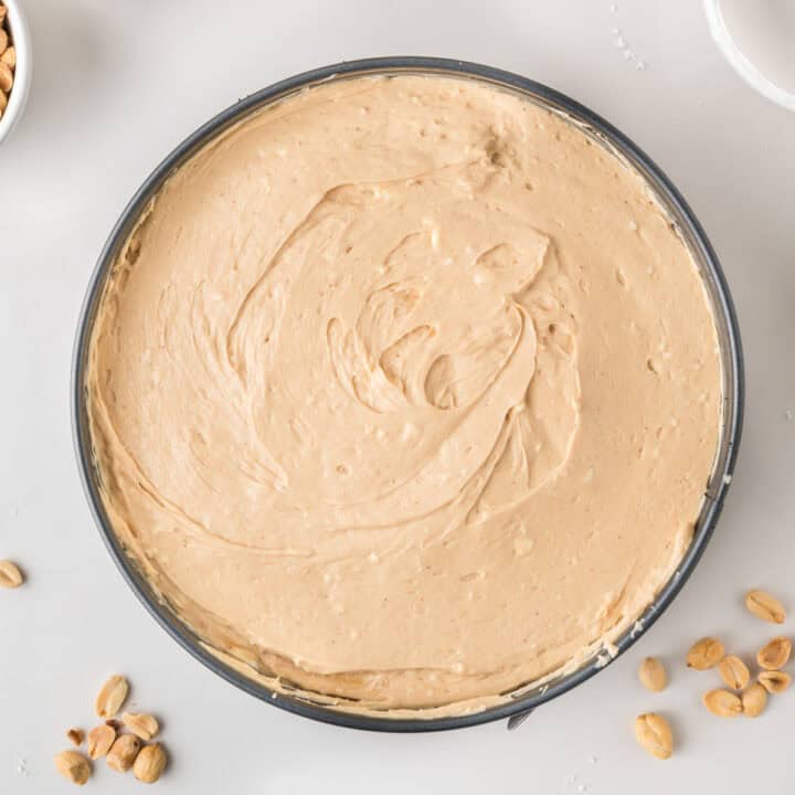 A round cake pan filled with smooth, creamy peanut butter cheesecake batter sits on a white surface, with scattered peanuts nearby.