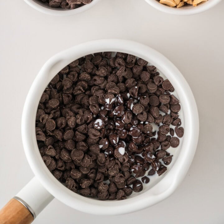 A white saucepan filled with chocolate chips and a small amount of melted chocolate sits on a white surface, with two bowls containing ingredients partially visible at the top of the image.