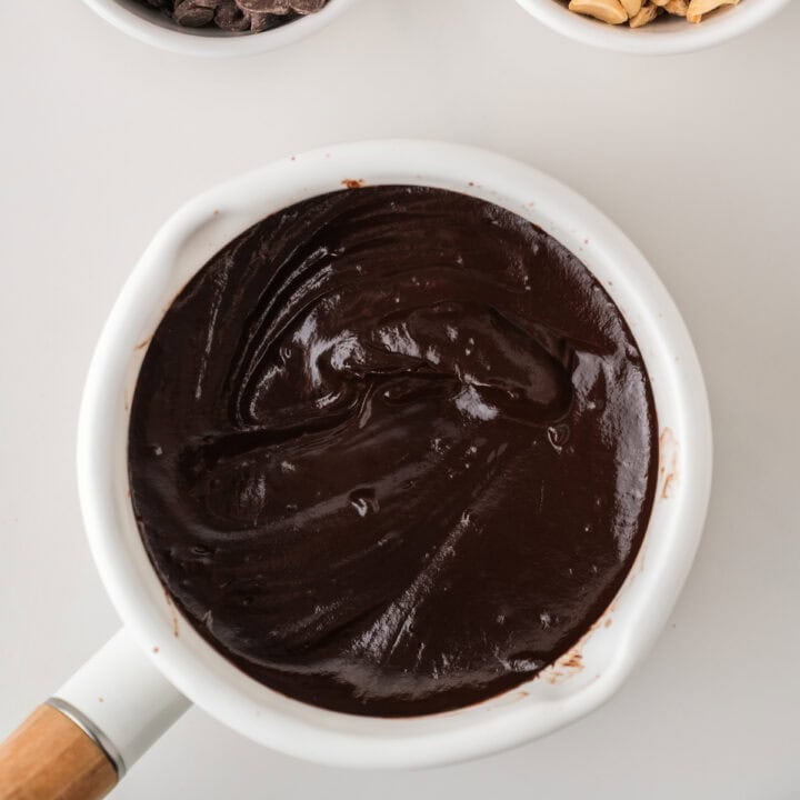 A white saucepan filled with thick, glossy melted chocolate sits on a white surface, with part of its wooden handle visible and small bowls containing chocolate chips and nuts nearby.